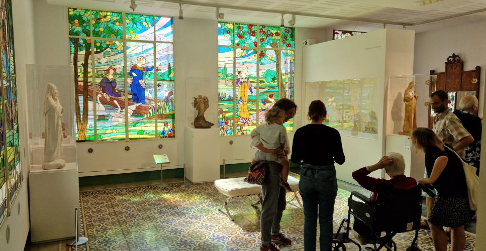 Visitants a la sala modernista del Museu d’Art de Cerdanyola observant els vitralls “Les dames de Cerdanyola”, amb escenes de figures femenines en un llac envoltat d’arbres fruiters i colors vius.