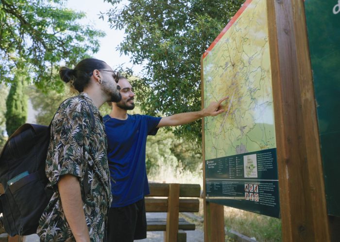 Informació turística al Parc Natural de Sant Llorenç de munt i l'Obac Dos nois joves vestits d’esport mirant un plafó amb el mapa de rutes al Parc Natural de Sant Llorenç del Munt i l’Obac.